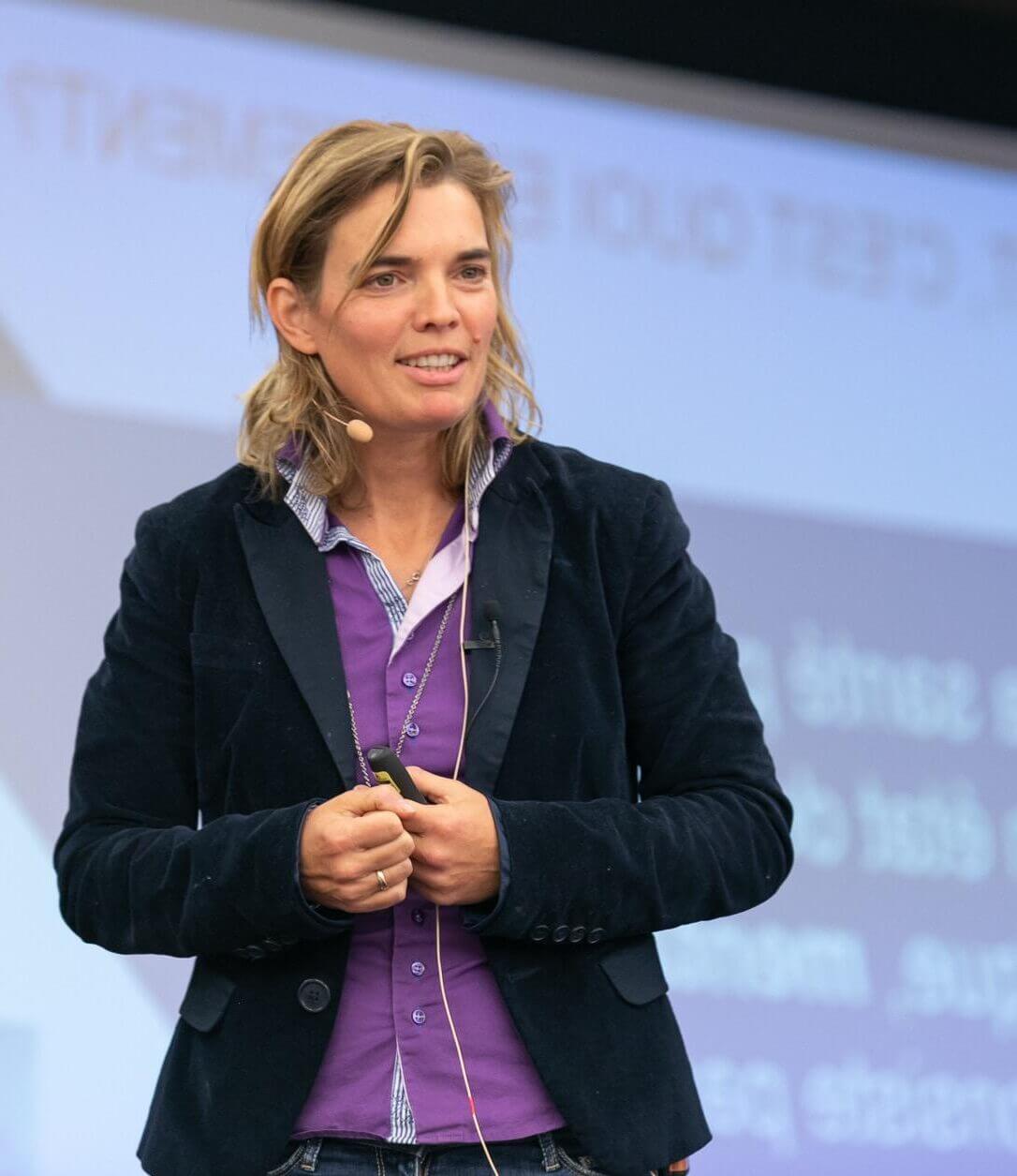 Photo de Caroline Heol, femme blonde aux cheveux mi-longs, elle porte une chemise violette et une veste blaser noir.