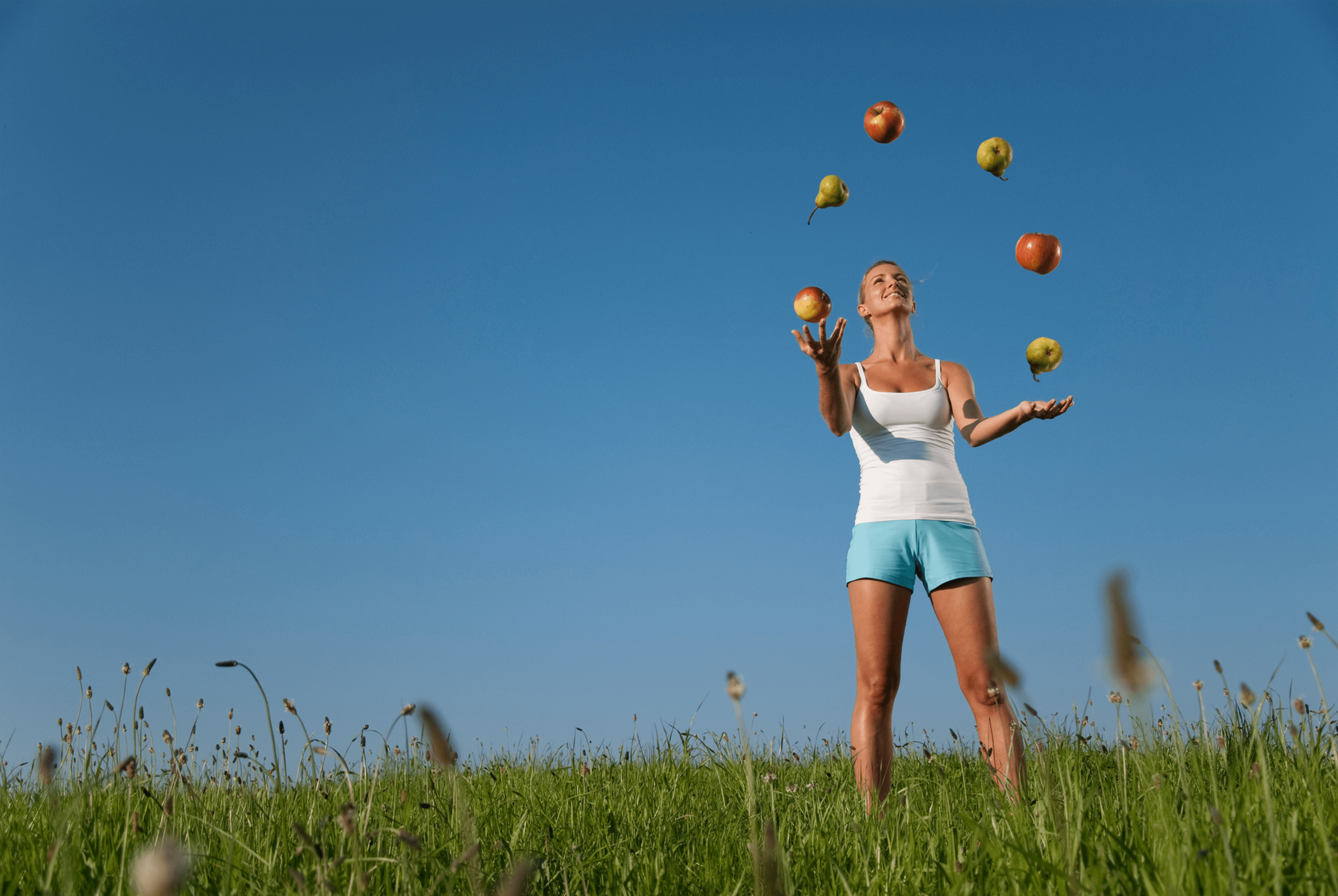 Photo d'une femme blonde en débardeur blanc et short bleu clair qui jongle avec des fruits. Elle est debout dans l'herbe.